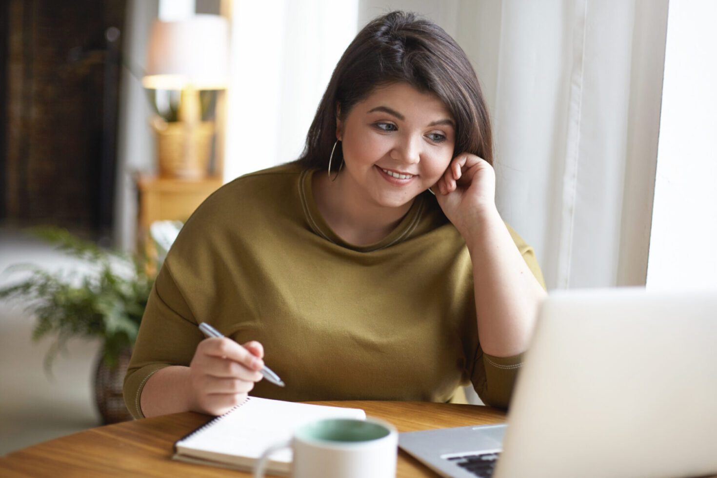 Indoor image of cheerful overweight student girl working on course paper, usign wireless internet connection on generic portable computer, making notes in copybook and smiling. Gadgets and education