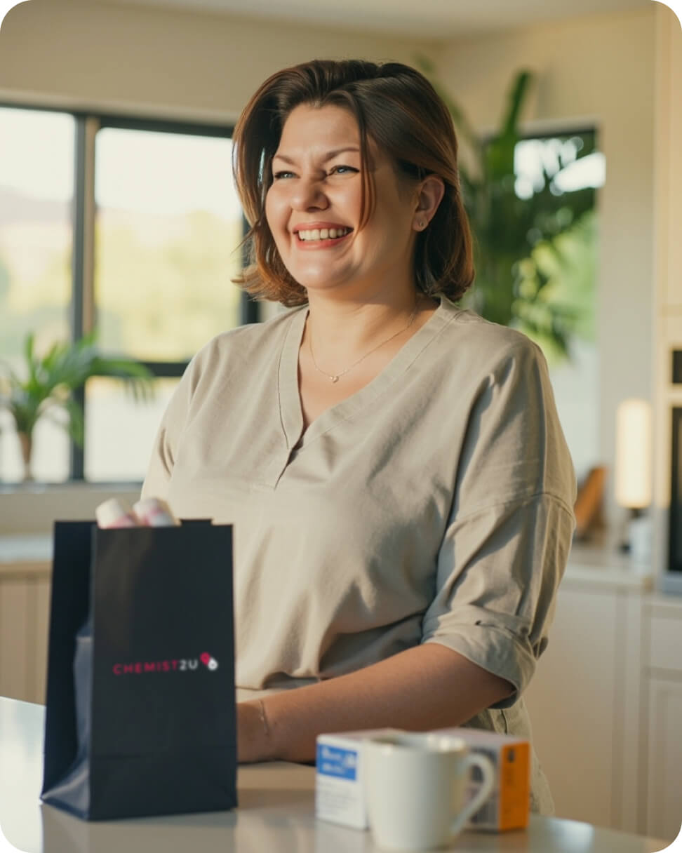A woman stands in her kitchen, smiling as she unpacks a navy Chemist2U bag filled with home-delivered medicines, surrounded by bright, modern countertops and natural light.